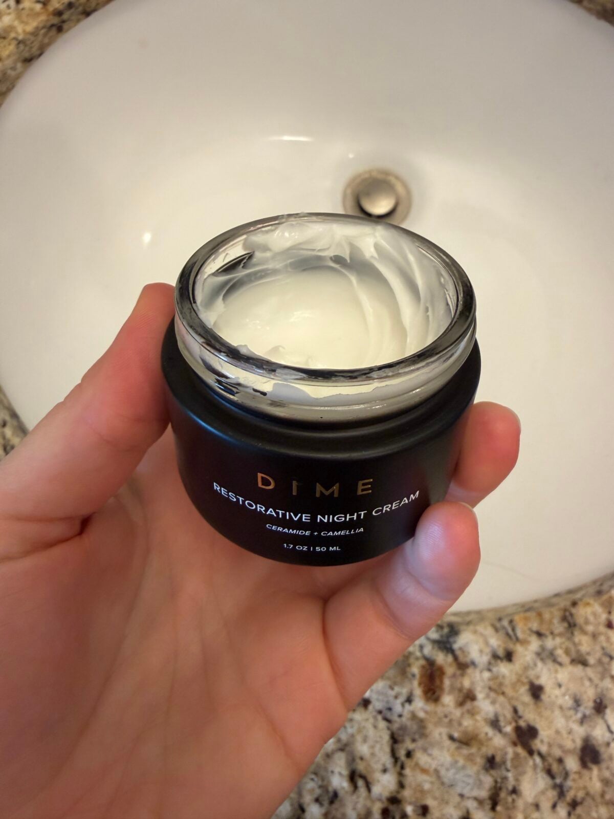 A hand holds an open jar of DIME Restorative Night Cream containing white cream, with a bathroom sink and granite countertop in the background.