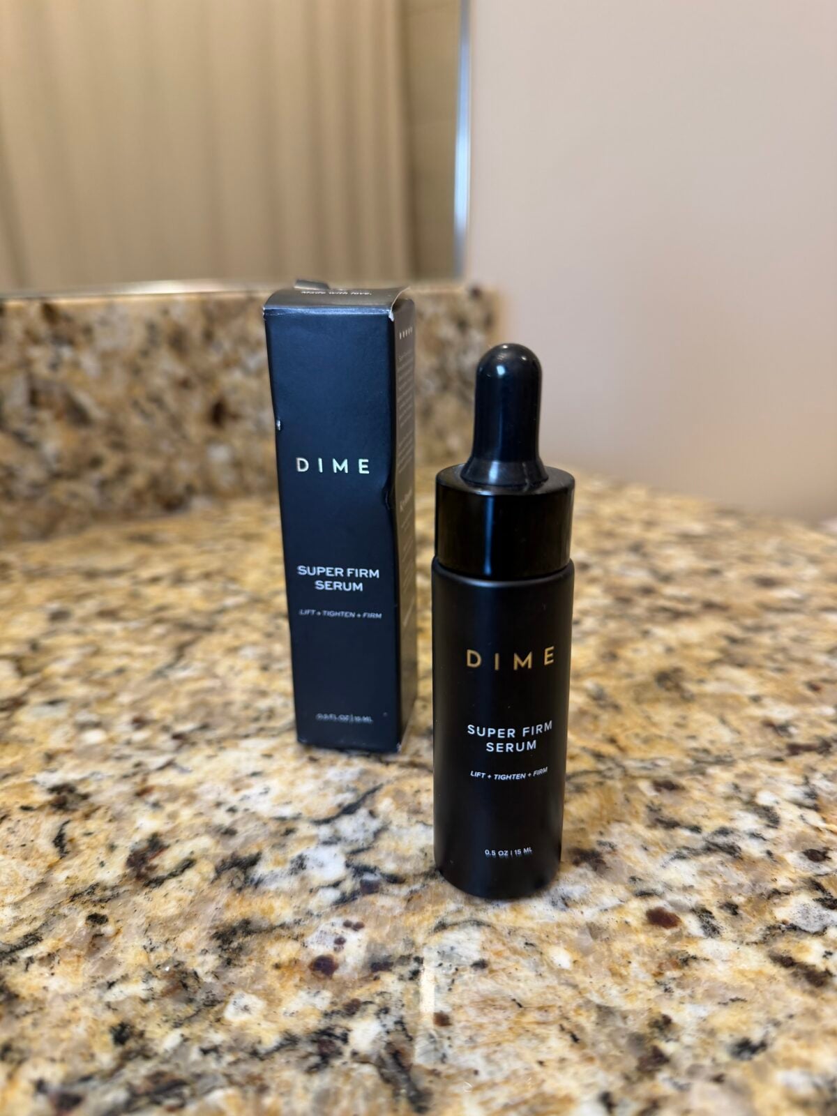 A bottle of DIME Super Firm Serum with a dropper is placed on a granite bathroom countertop. The product&rsquo;s box stands upright behind the bottle, and a blurred mirror is visible in the background.