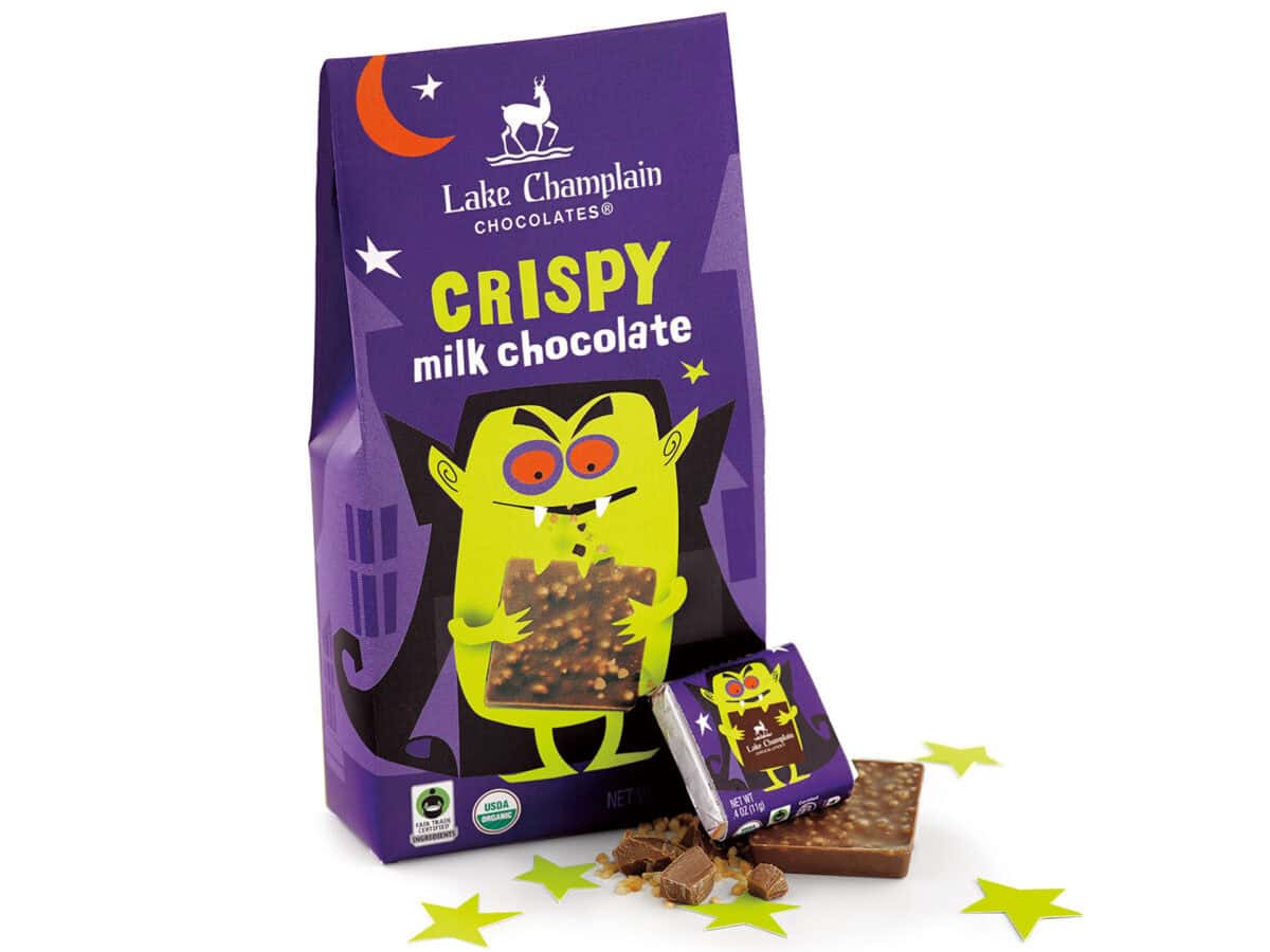 A purple bag of Lake Champlain Chocolates Crispy Milk Chocolate is shown, featuring a cartoon vampire illustration and a broken-off piece of chocolate on a white background with yellow stars.
