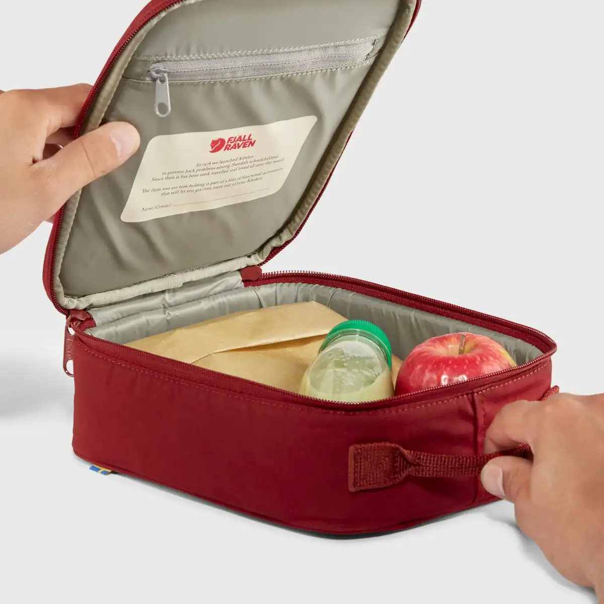 The maroon Kanken Mini Cooler by Fj&auml;llr&auml;ven, with a drink and apple inside, fully opened.