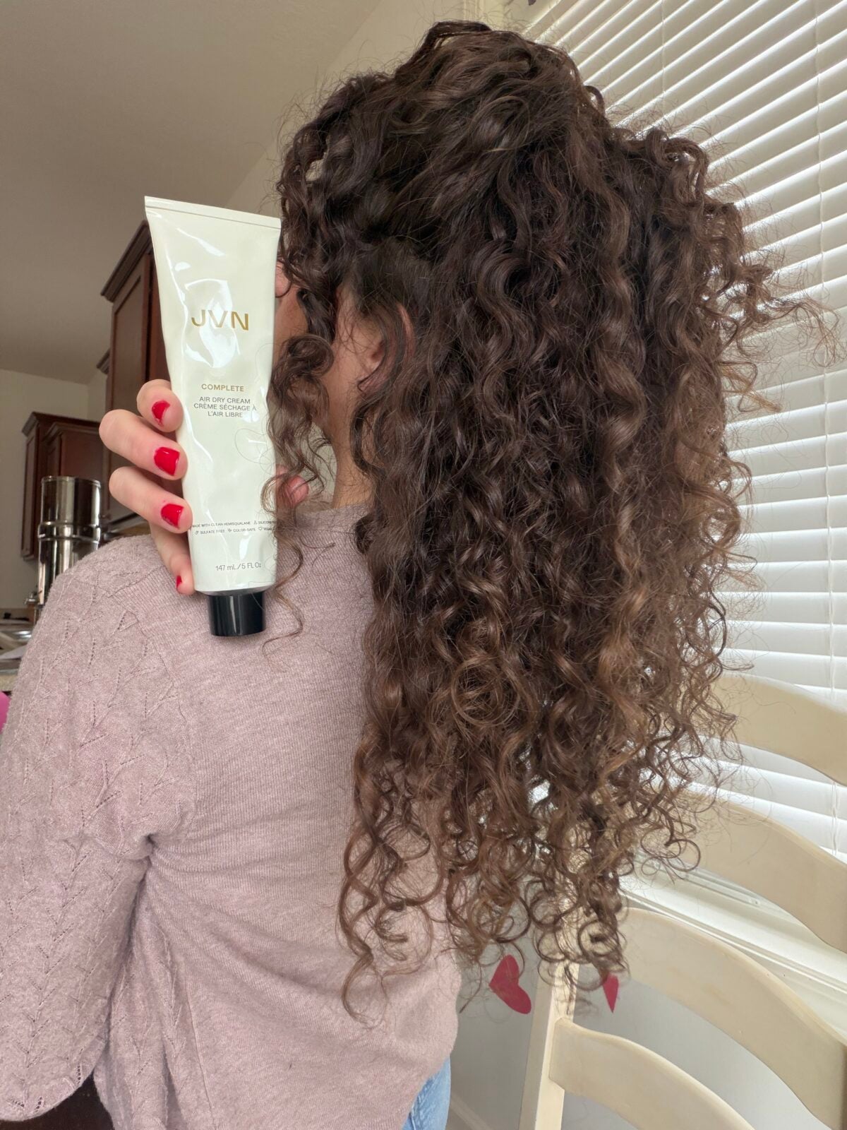 A person with curly hair holds a tube of JVN Complete Air Dry Cream.