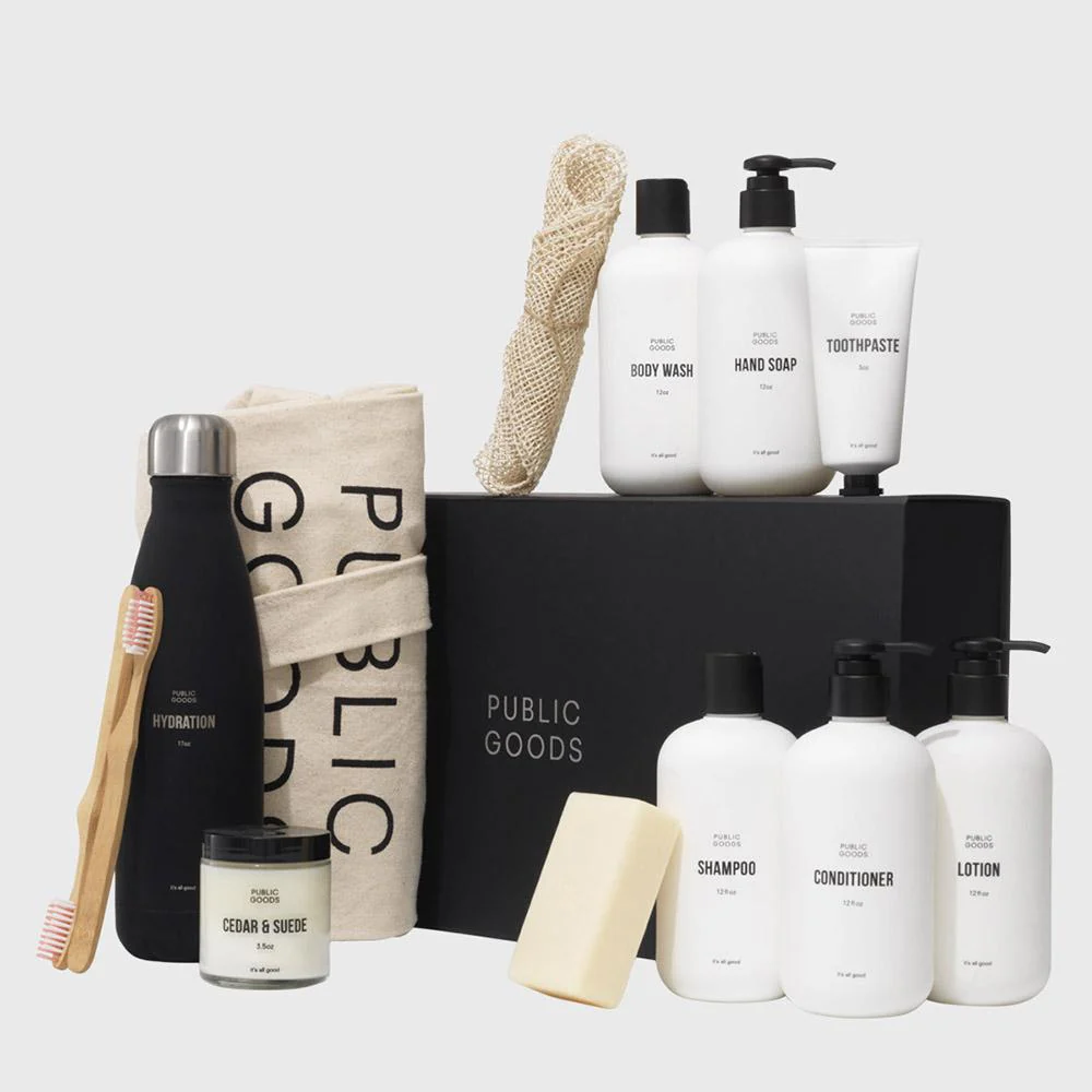 The photo displays a variety of Public Goods personal care and home products, including a black water bottle, a canvas tote bag, bar soap, and various white bottles of shampoo and lotion, arranged around a black "PUBLIC GOODS" box.