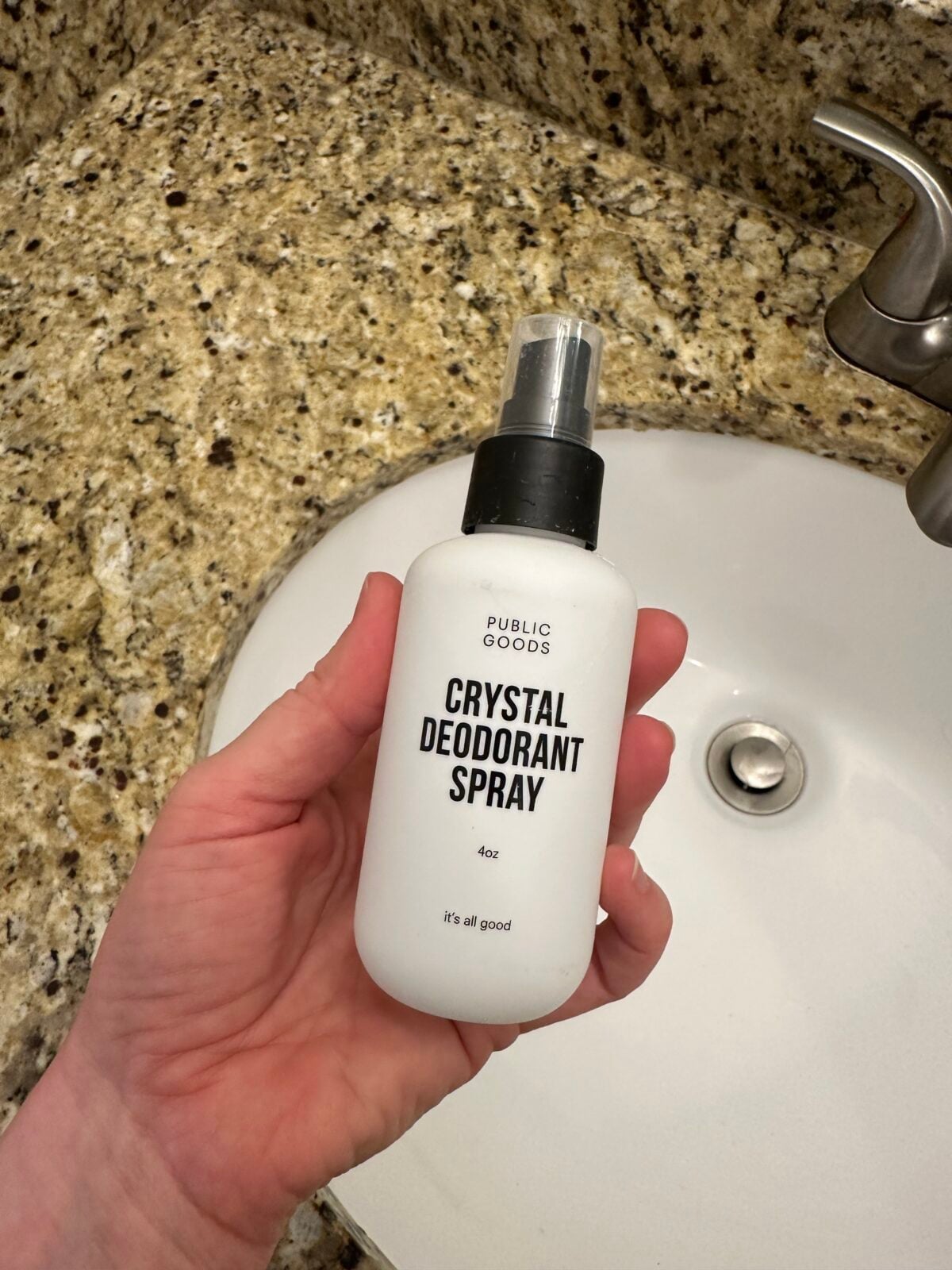 Hand holding Public Goods crystal deodorant spray bottle, 4 oz, over bathroom sink with granite counter