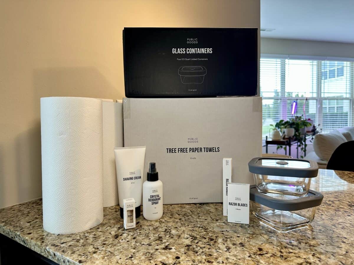 A collection of Public Goods products displayed on a granite kitchen counter, including tree-free paper towels, glass food containers with gray lids, shaving cream, crystal deodorant spray, razor blades, and a razor handle. Large boxes labeled &ldquo;Glass Containers&rdquo; and &ldquo;Tree Free Paper Towels&rdquo; are stacked in the background, with natural light and houseplants visible through nearby windows.