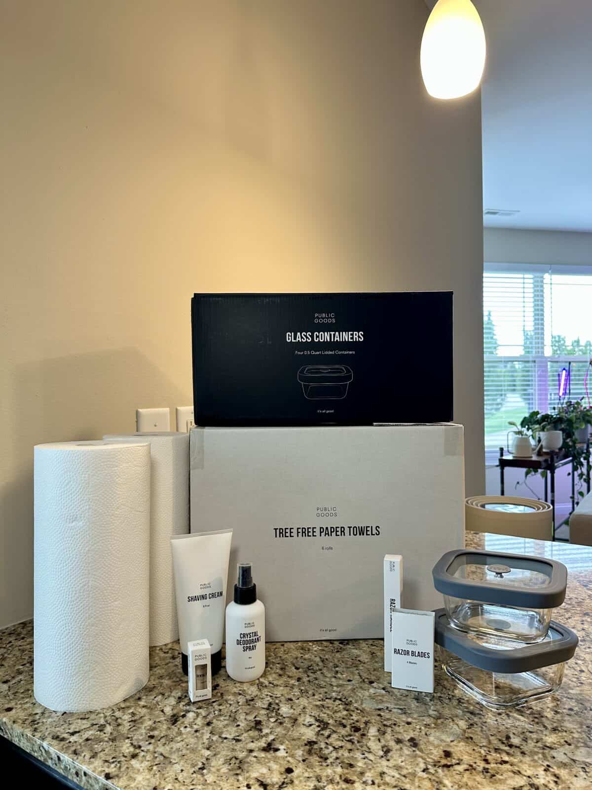 A collection of Public Goods products displayed on a granite kitchen counter, including tree-free paper towels, glass food containers with gray lids, shaving cream, crystal deodorant spray, razor blades, and a razor handle. Large boxes labeled “Glass Containers” and “Tree Free Paper Towels” are stacked in the background, with natural light and houseplants visible through nearby windows.