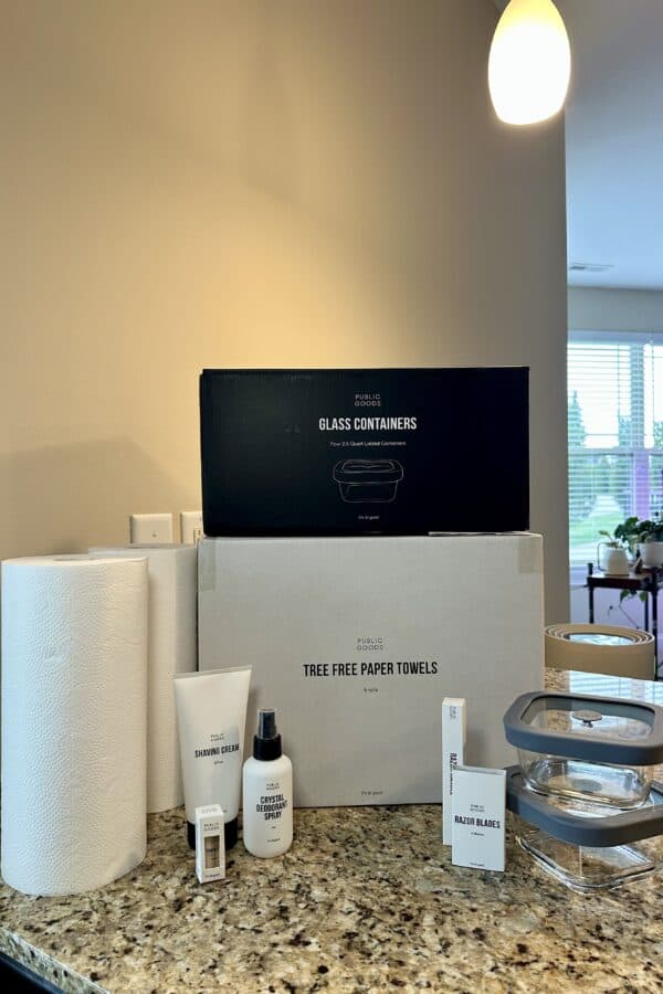 A collection of Public Goods products displayed on a granite kitchen counter, including tree-free paper towels, glass food containers with gray lids, shaving cream, crystal deodorant spray, razor blades, and a razor handle. Large boxes labeled “Glass Containers” and “Tree Free Paper Towels” are stacked in the background, with natural light and houseplants visible through nearby windows.