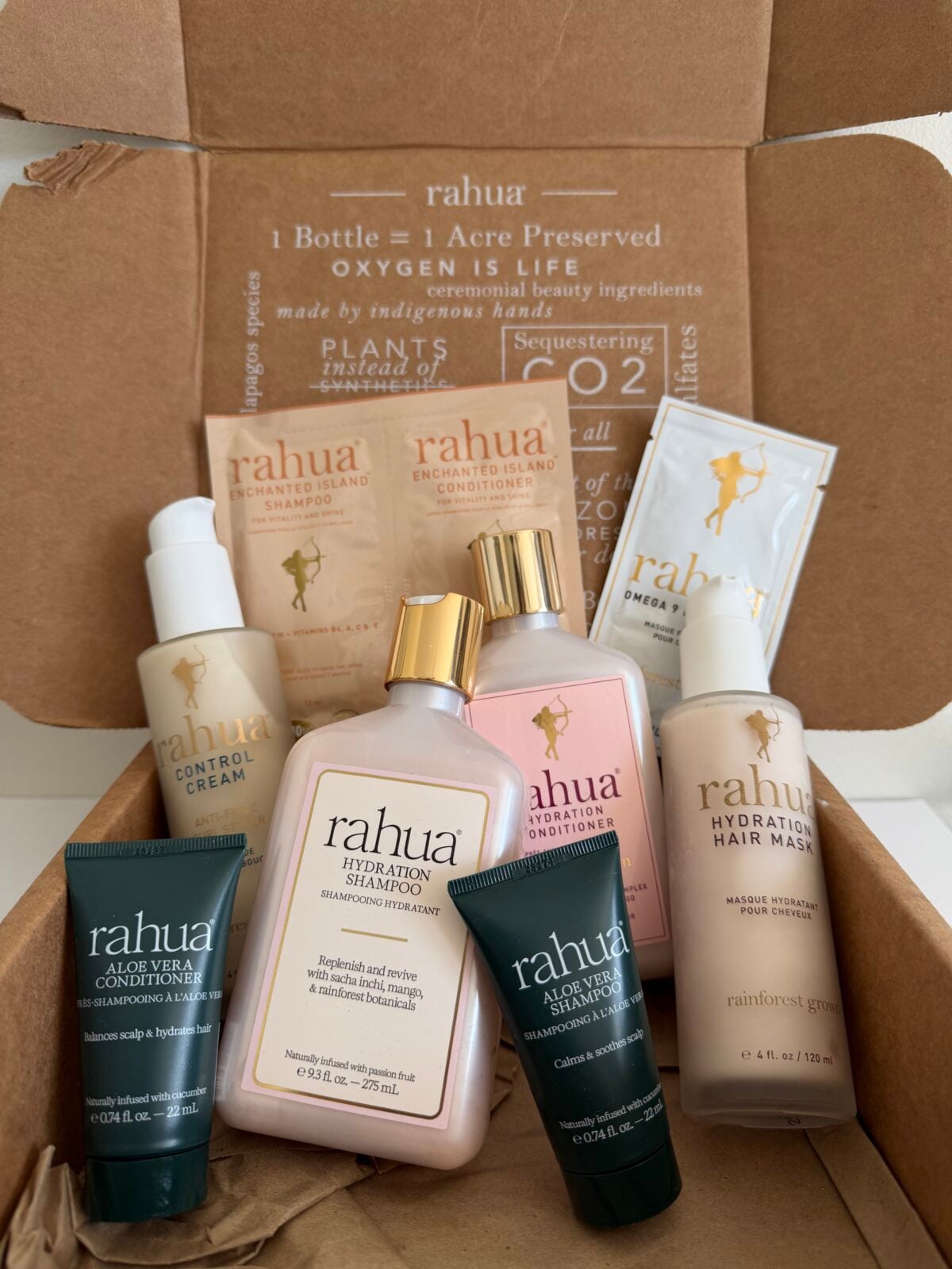 A cardboard shipping box containing an assortment of Rahua hair care products, including full-size bottles of Hydration Shampoo and Hydration Hair Mask, smaller tubes of Aloe Vera shampoo and conditioner, and several sample packets.