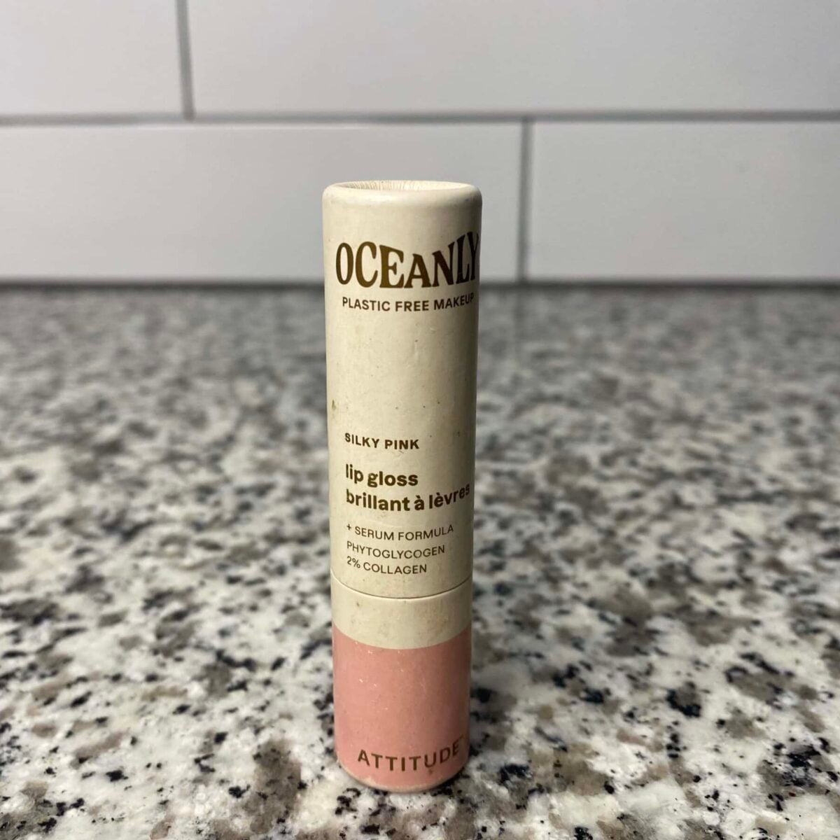 The photo displays a single tube of Oceanly "Silky Pink" Lip Gloss by the brand Attitude, which features a plastic-free paper tube and a serum formula containing phytoglycogen and collagen, standing upright on a speckled granite countertop.