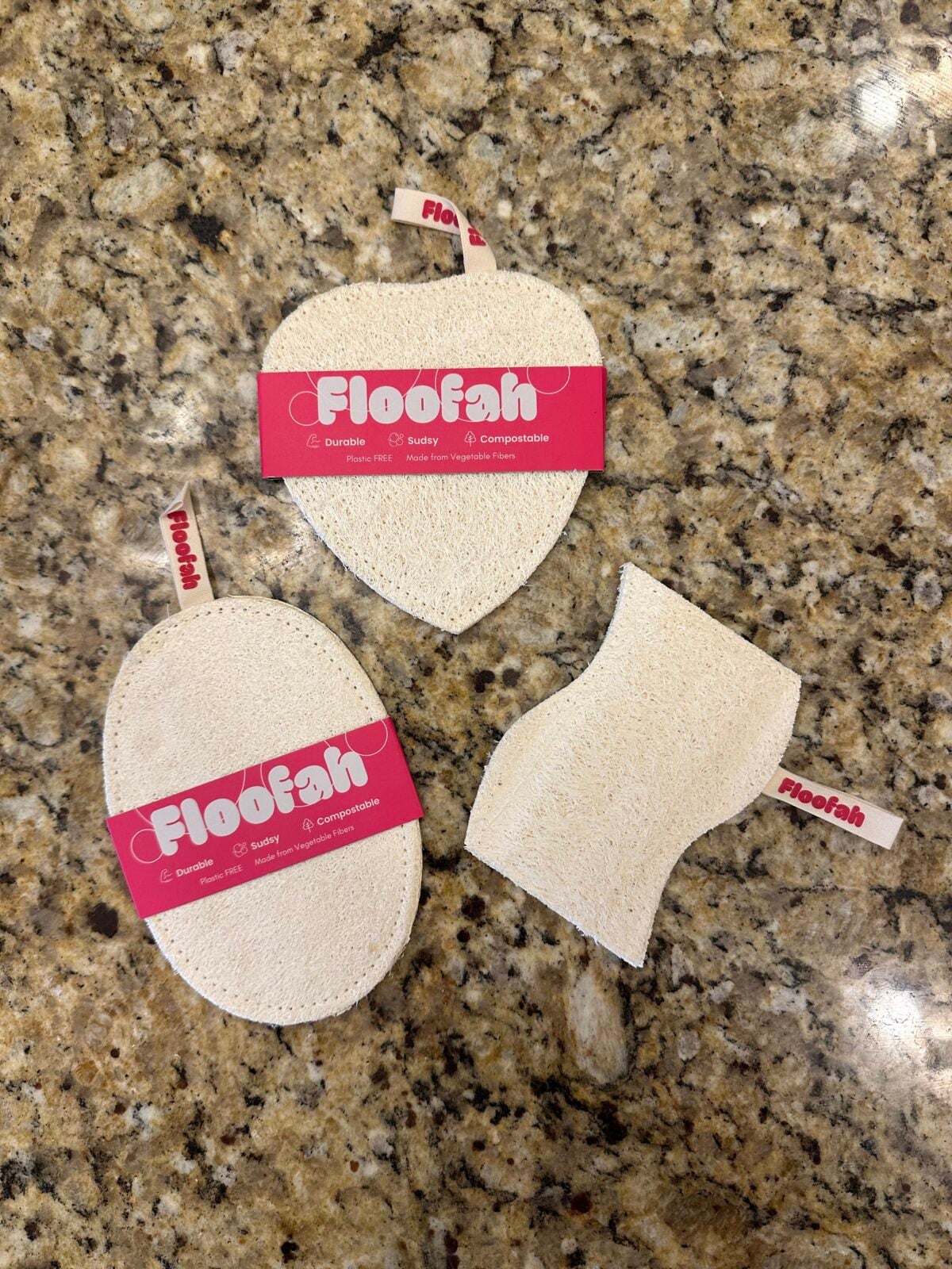 Three natural loofah sponges in different shapes—heart, oval, and curved—each with a red Floofah label, laid out on a granite countertop.