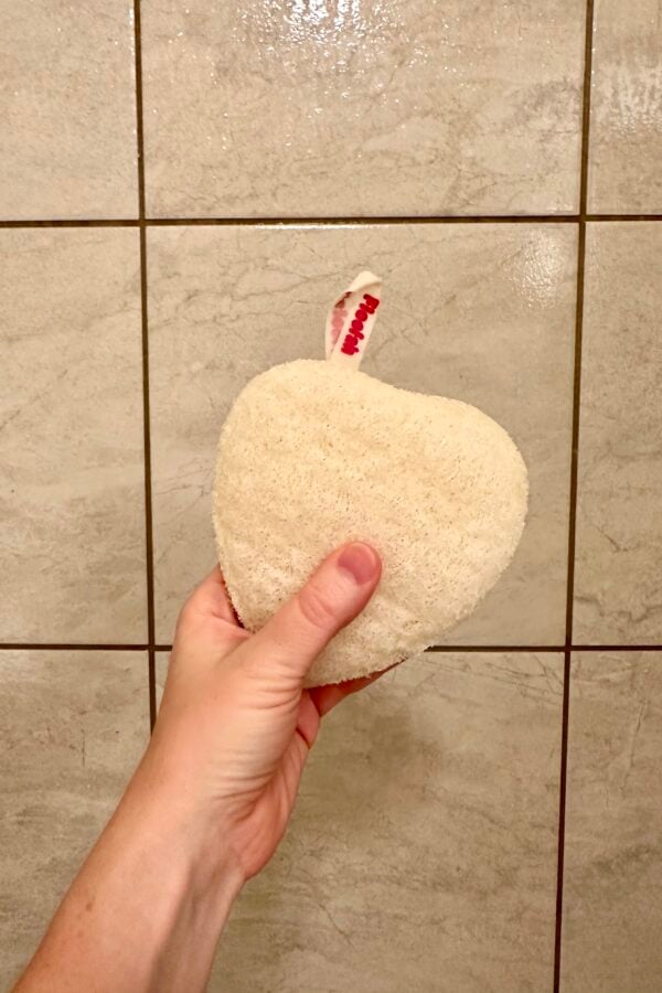 Hand holding a heart-shaped natural loofah sponge with a small red-and-white hanging loop against a tiled shower wall.