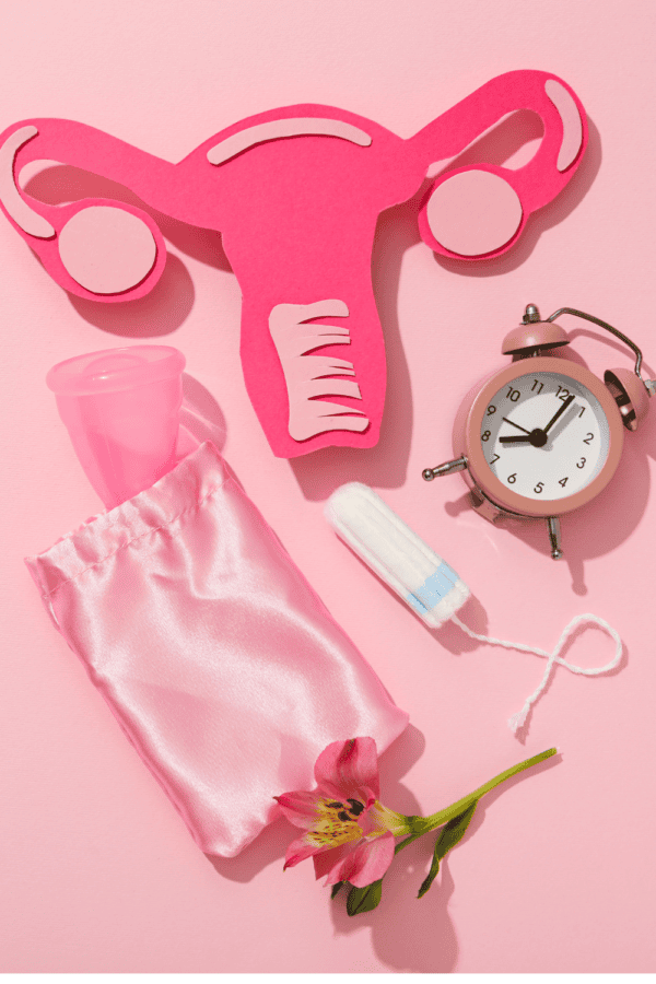 A pink background with a paper cutout of a uterus, a tampon, a flower, a menstrual cup, and a clock