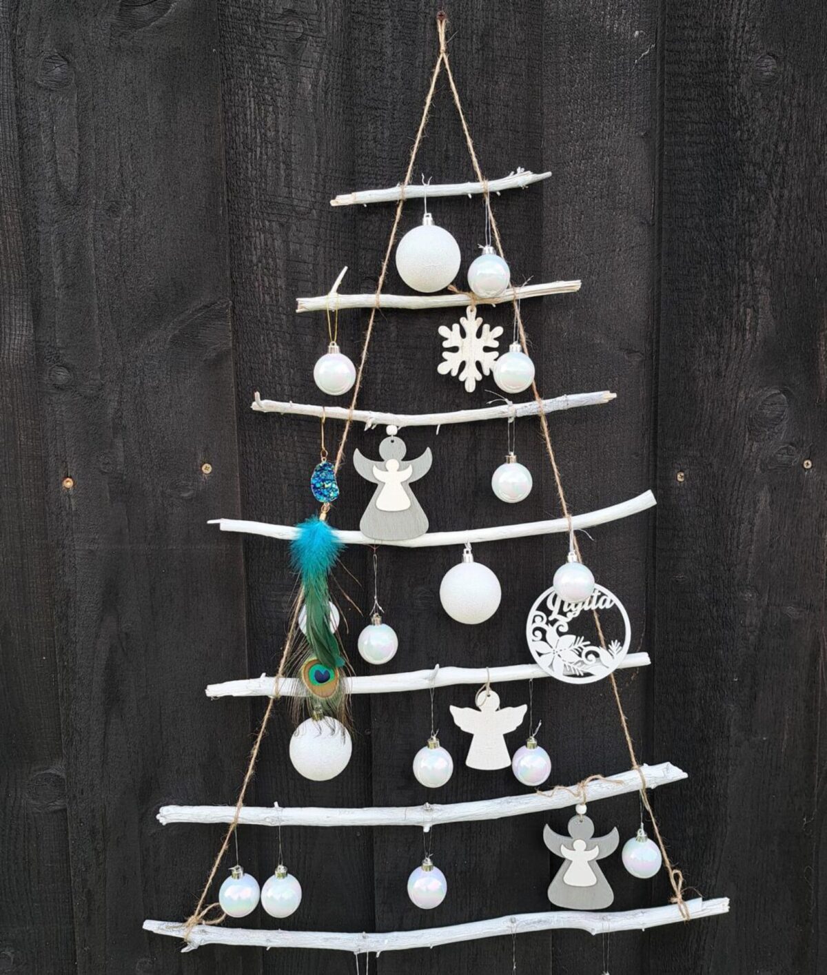 Wall-hanging Christmas tree made from white painted branches and twine, decorated with white ornaments, wooden angels, a snowflake, and a peacock feather.