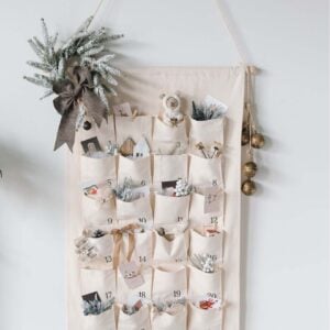 Neutral fabric advent calendar with numbered pockets, filled with small holiday items and greenery.
