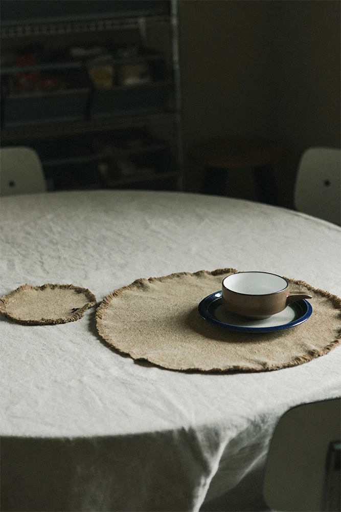 Neutral round jute placemats on a linen-covered table with ceramic dishes in a cozy, dim kitchen setting.