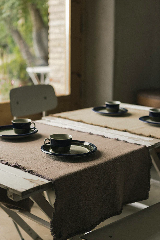 Natural woven table runners on a rustic wooden table set with ceramic plates and cups in a bright dining space.