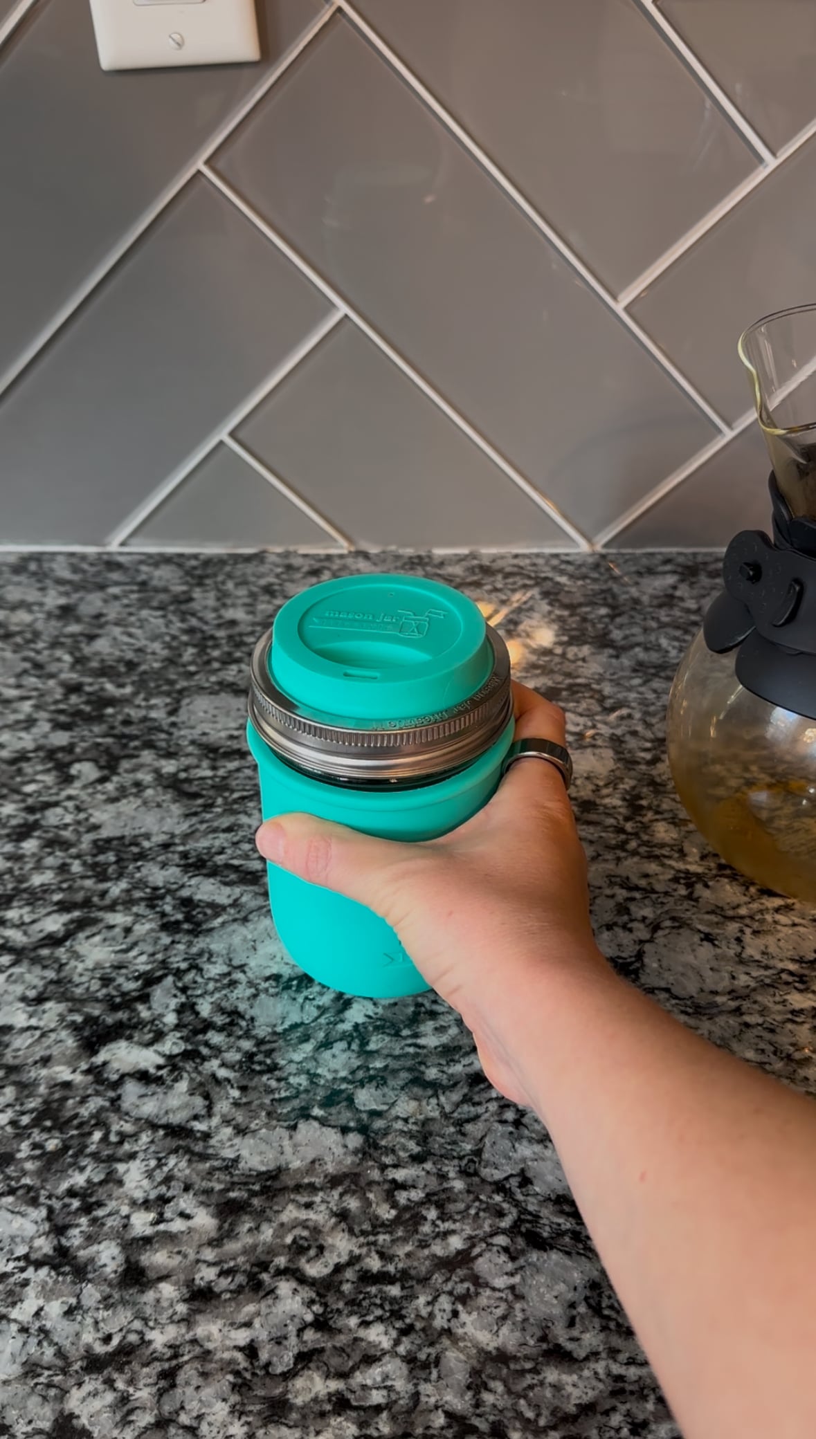 Hand holding a turquoise silicone-sleeved mason jar with a matching reusable lid on a gray granite countertop.