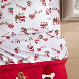 Bed with white Christmas bedding featuring Santa and reindeer print, layered with red sheets and a festive pillow.
