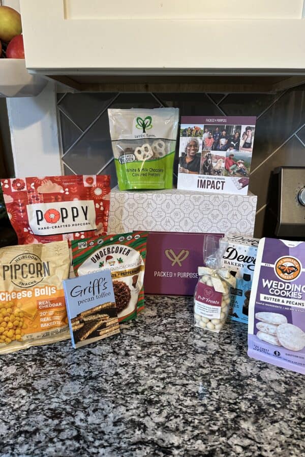 A collection of gourmet snacks and treats from a Packed with Purpose gift box, including cheese popcorn, pretzels, toffee, and cookies, are arranged on a dark kitchen countertop.