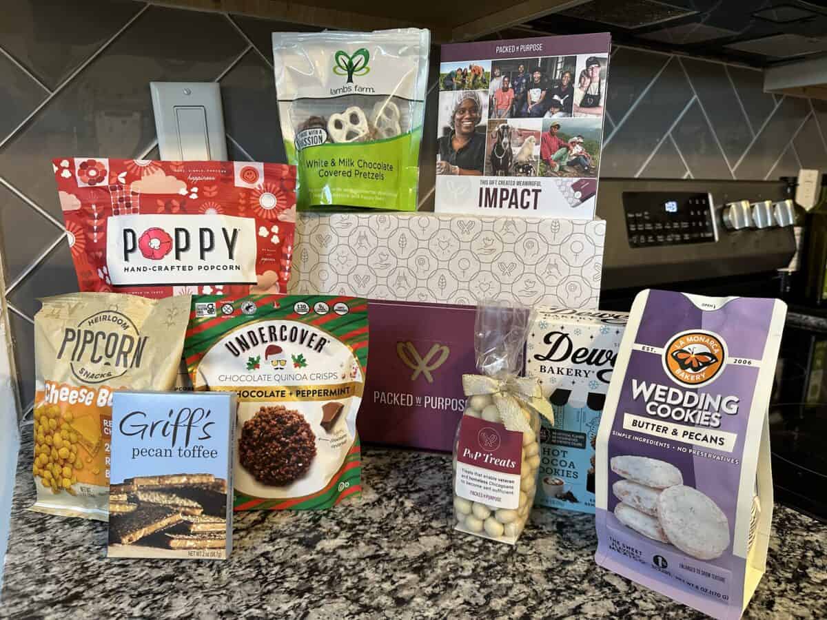 A collection of various gourmet snacks and treats from a Packed with Purpose gift box, including pretzels, popcorn, toffee, and cookies, are arranged on a dark kitchen countertop.