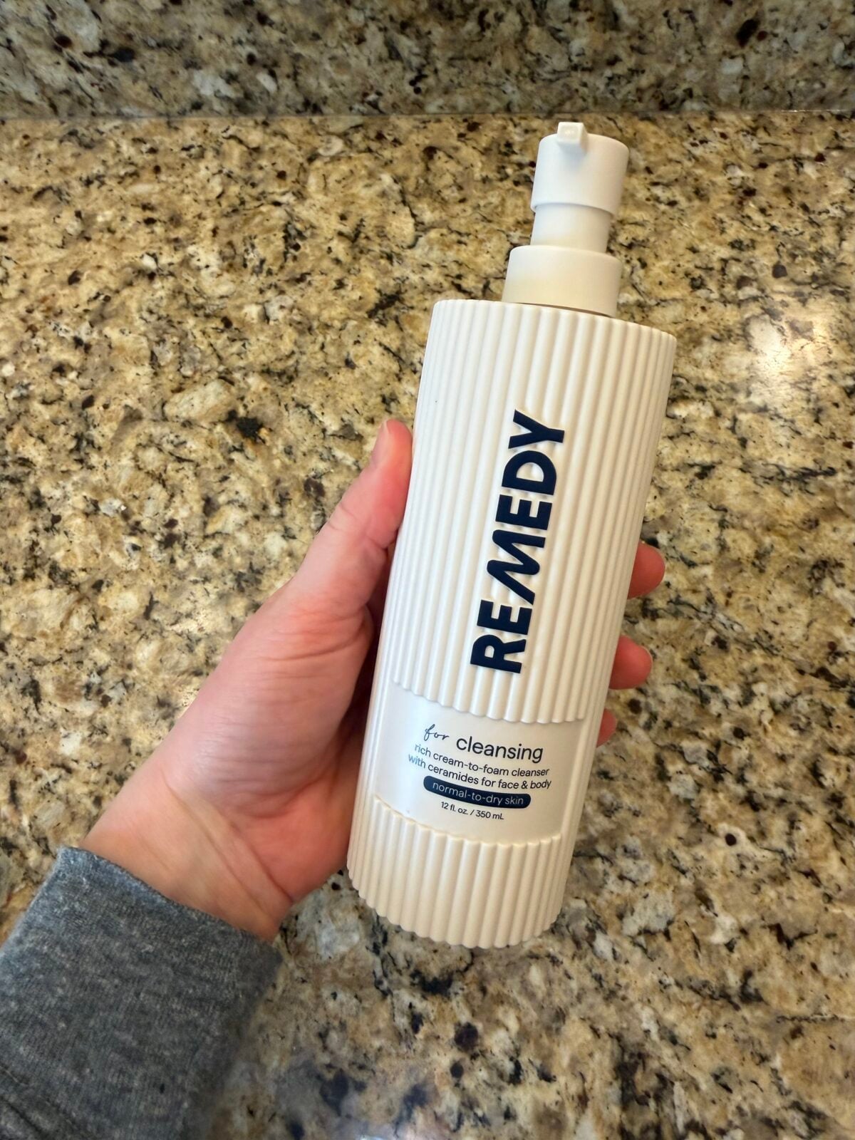 Hand holding Remedy brand cream-to-foam cleanser bottle with white ribbed packaging against granite countertop