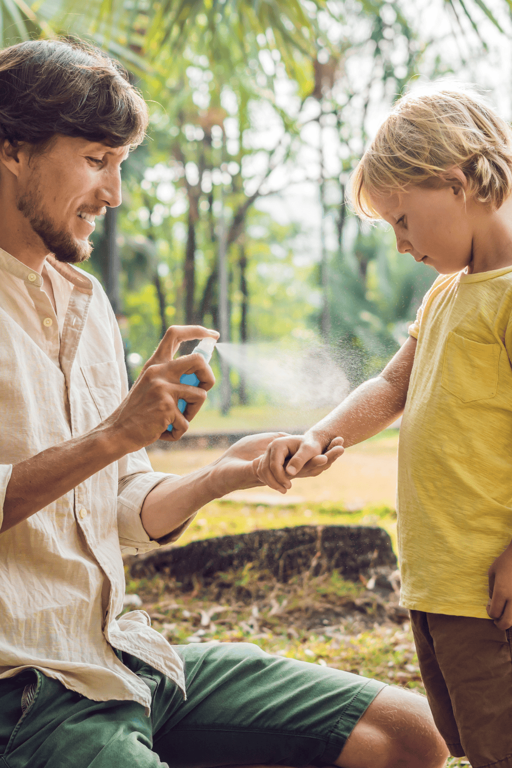 a parent and a child outside. the parent is kneeling down spraying insect repellent on the child's arm