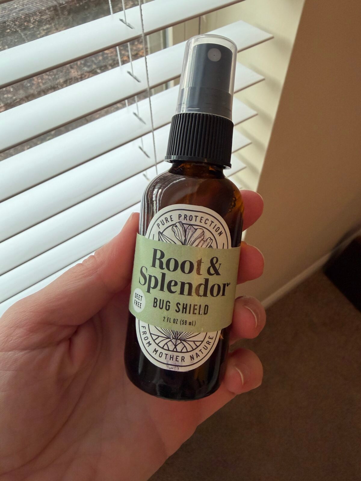 Hand holding Root & Splendor Bug Shield DEET-free natural bug spray in amber glass bottle, 2 fl oz, in front of window with blinds