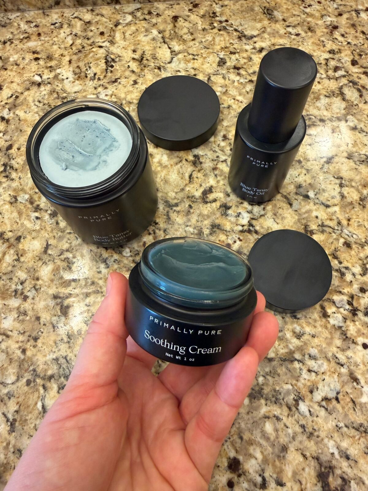 A hand holds an open jar labeled Primally Pure Soothing Cream with a blue-green cream inside. Two other opened skincare containers are on a granite countertop in the background.