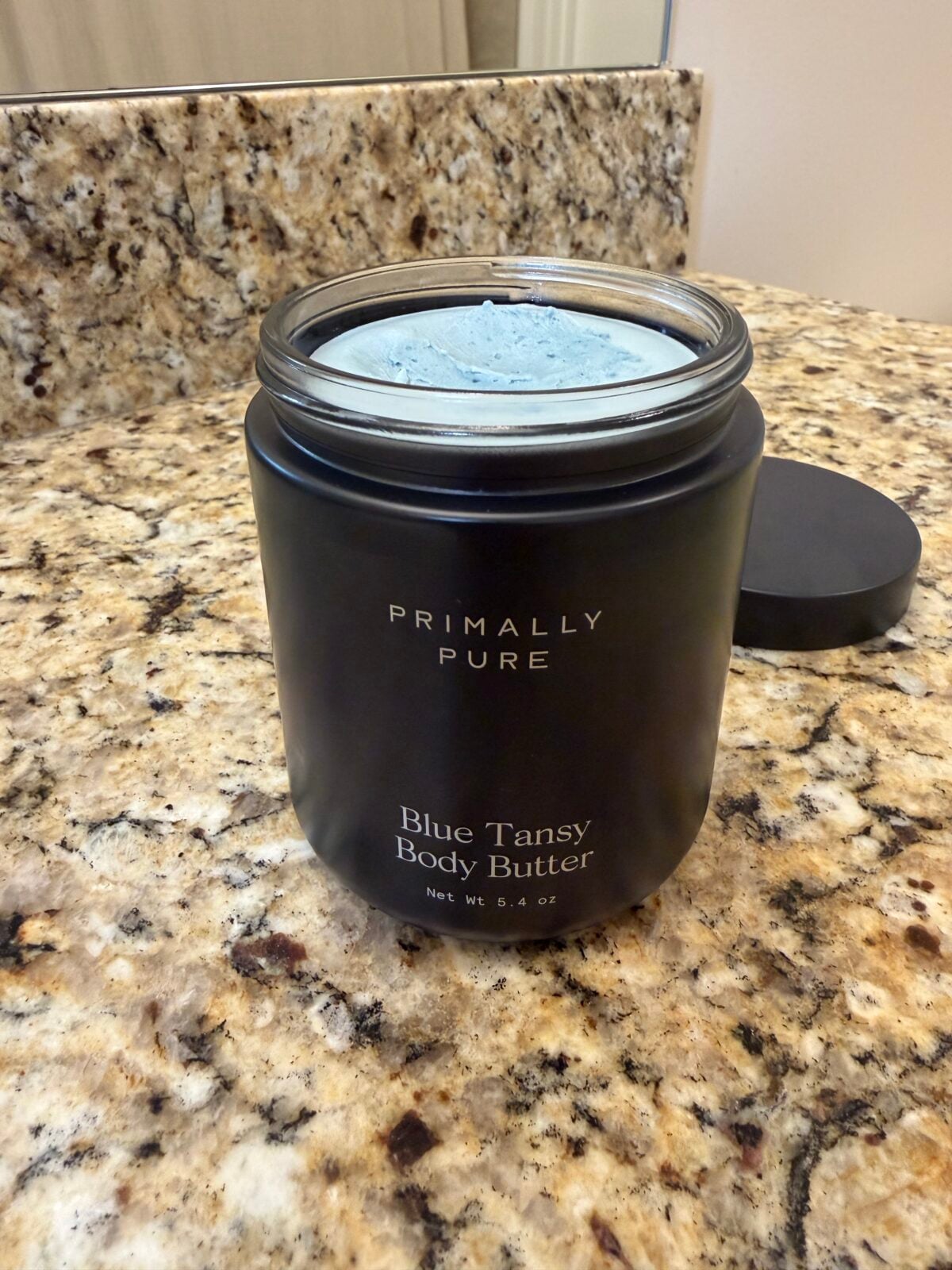A black jar labeled Primally Pure Blue Tansy Body Butter sits open on a granite bathroom countertop, showing pale blue body butter inside. The black lid is placed nearby.