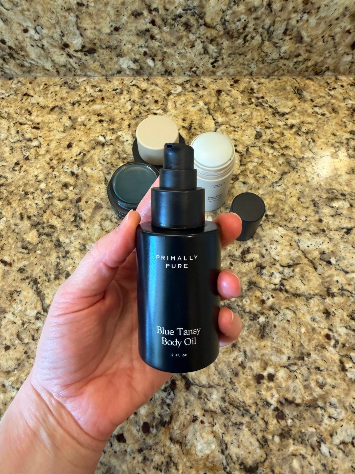 A hand holds a black bottle labeled Primally Pure Blue Tansy Body Oil above a granite countertop with other skincare containers in the background.