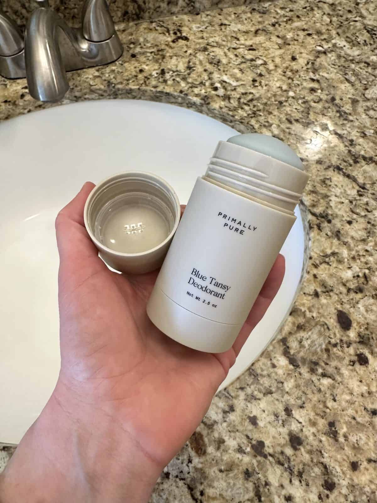 Hand holding an open Primally Pure Blue Tansy Deodorant over a bathroom sink with a granite countertop.