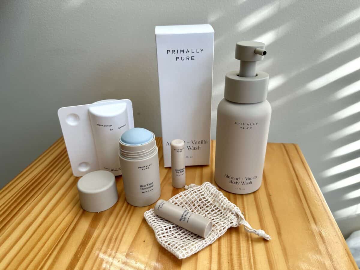 Primally Pure skincare products arranged on a wooden surface, including a Blue Tansy deodorant, Almond + Vanilla body wash, lip balm, and a small mesh bag, with soft natural lighting.