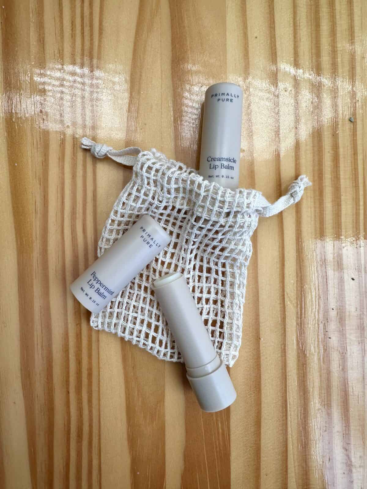 Primally Pure lip balms, including Creamsicle and Peppermint flavors, displayed on a wooden surface with a small mesh bag.