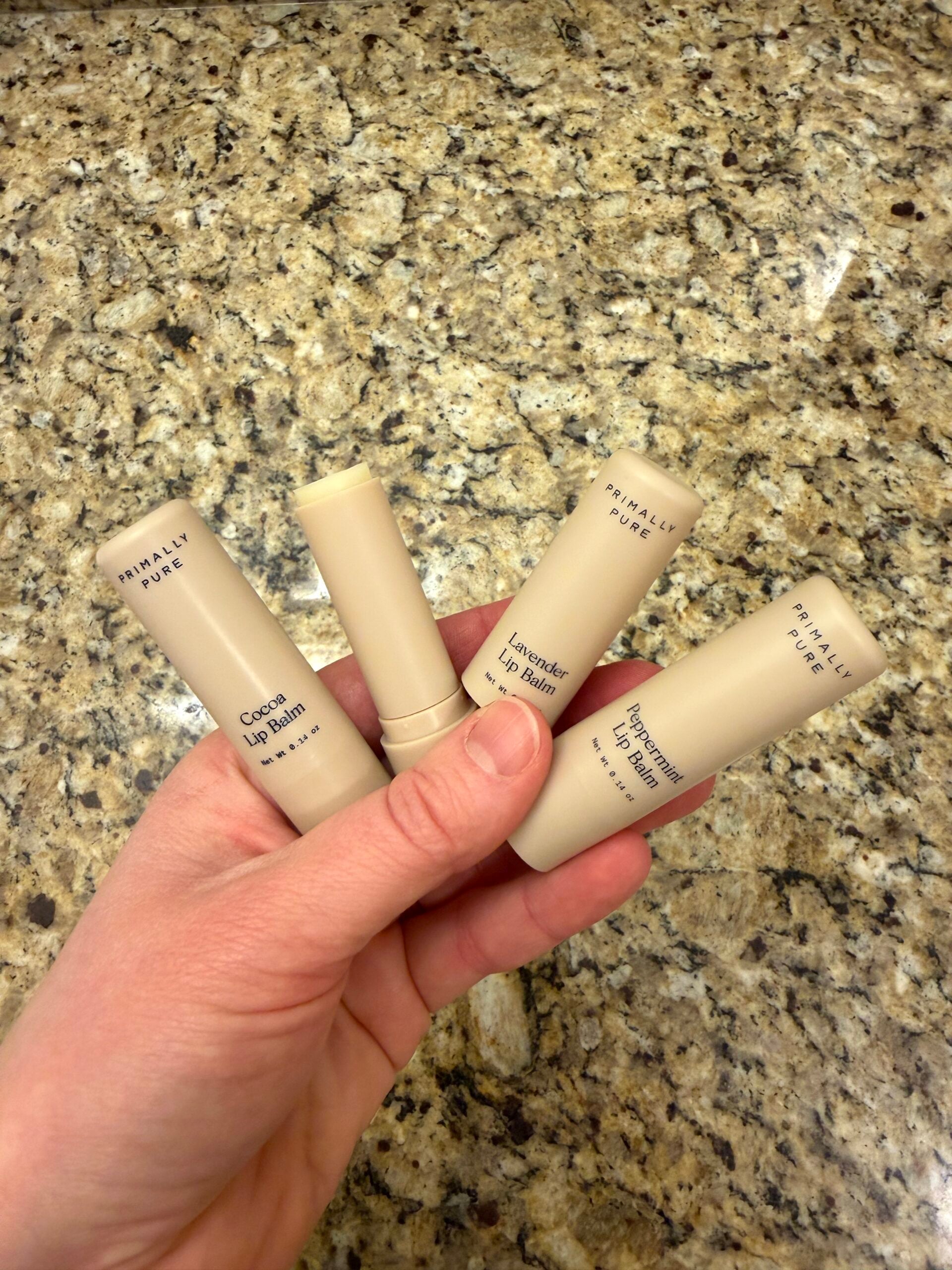 Hand holding three Primally Pure lip balm tubes in Cocoa, Lavender, and Peppermint flavors on granite countertop