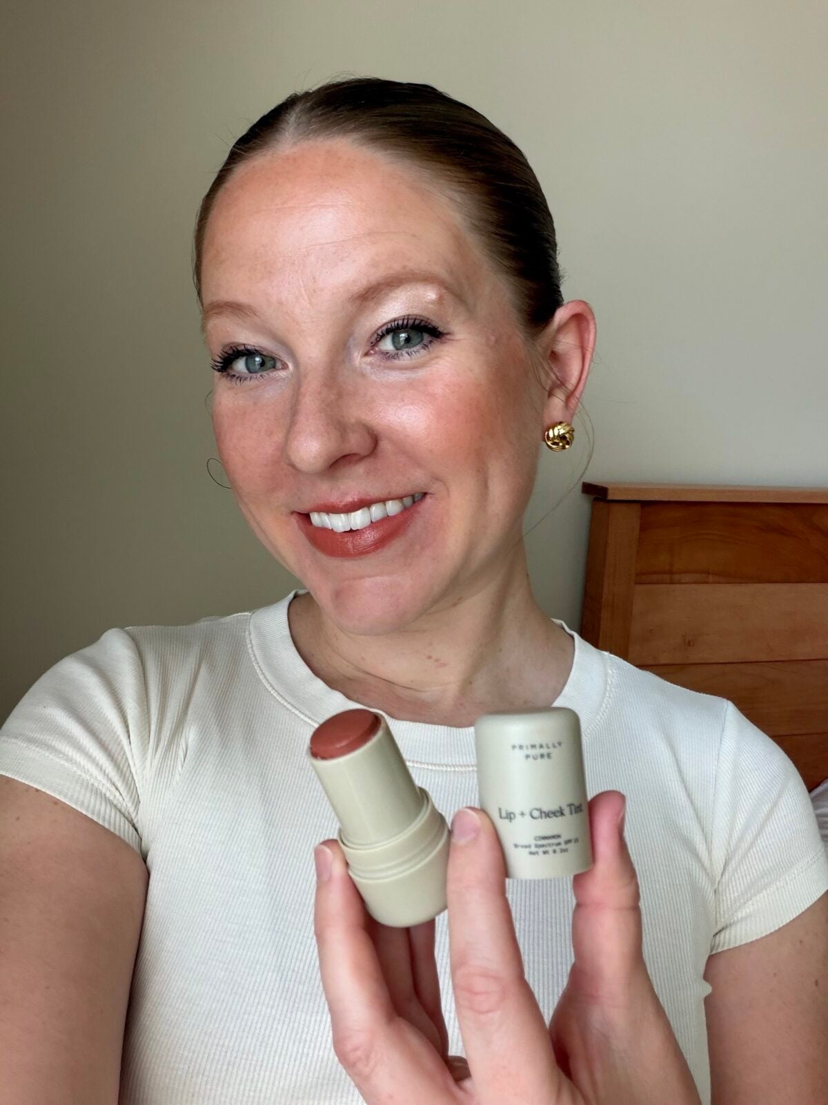 A woman with a light complexion smiles while holding a tube of lip and cheek tint with the cap off. She is wearing a white shirt and gold earrings, posing indoors near a wooden headboard.