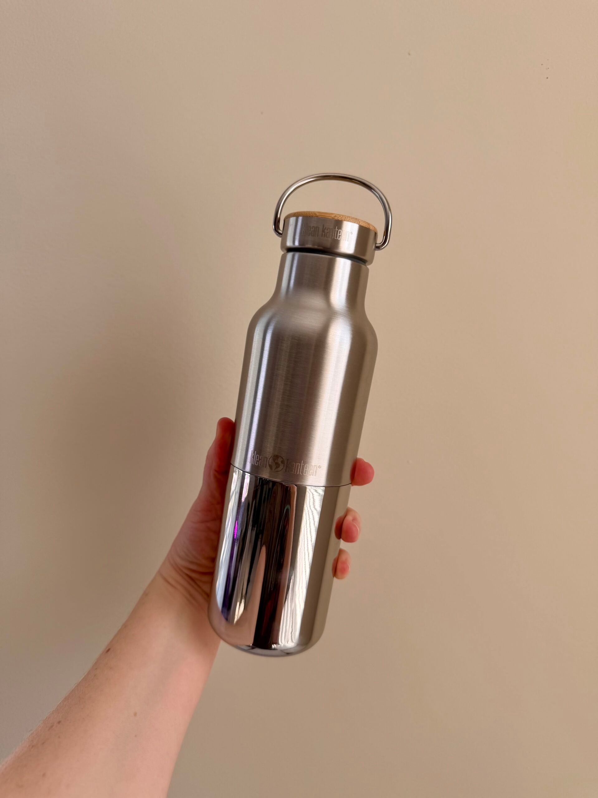 A hand holds a stainless steel reusable water bottle with a handle on the lid against a plain beige background.