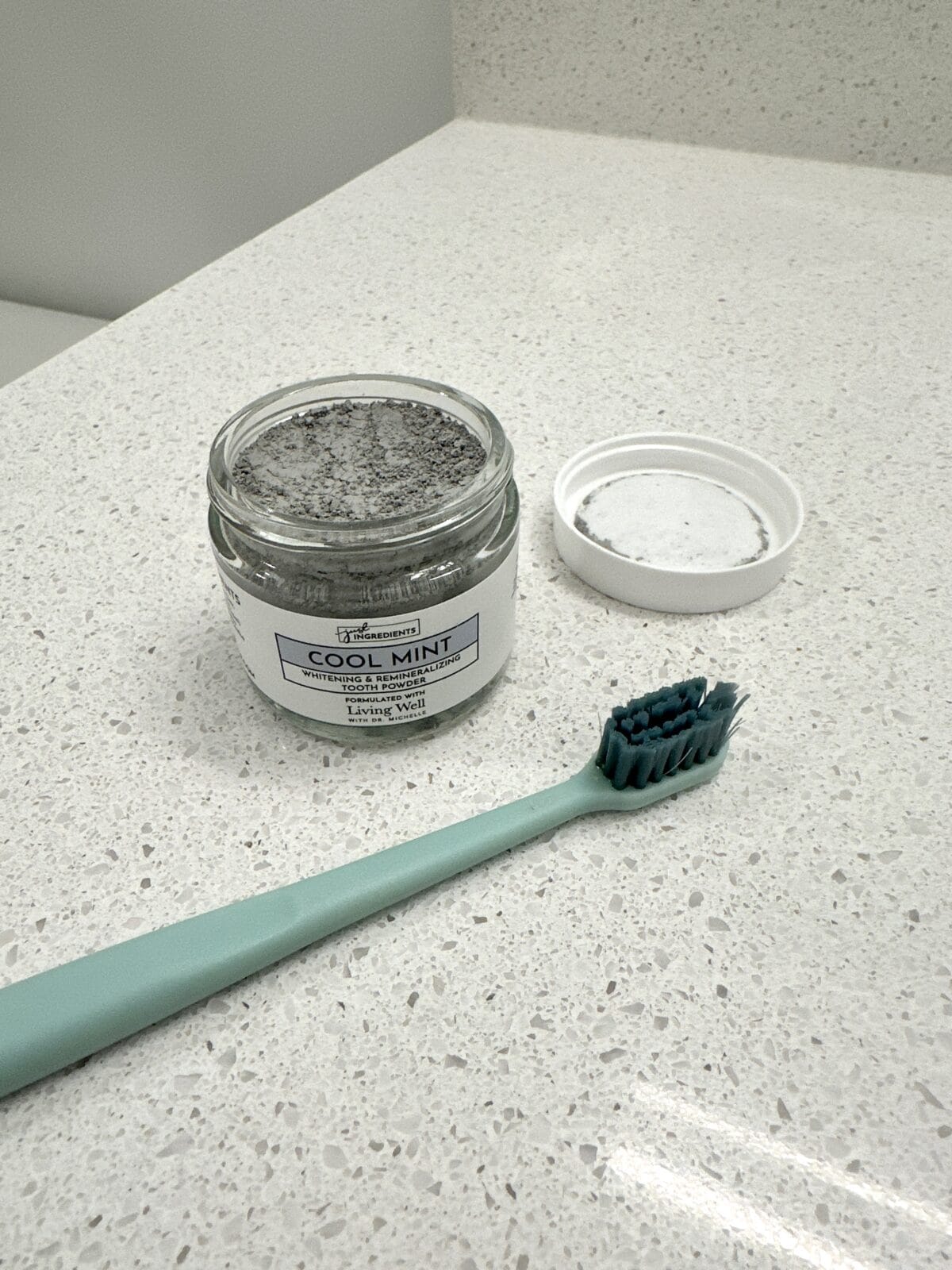 A glass jar of Just Ingredients Cool Mint Whitening & Remineralizing Tooth Powder, with a toothbrush beside it.