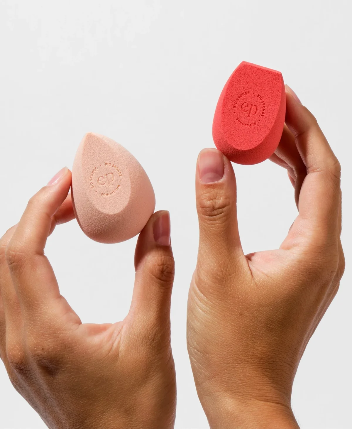 Two hands holding non-toxic makeup sponges from Ere Perez in peach and red colors, showcasing their unique shapes and embossed branding.