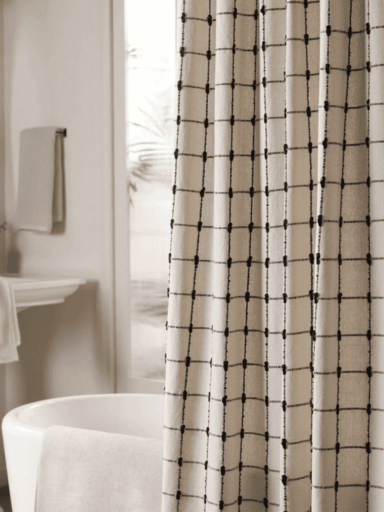 Beige shower curtain with a black grid pattern hanging beside a white bathtub in a bright, modern bathroom.