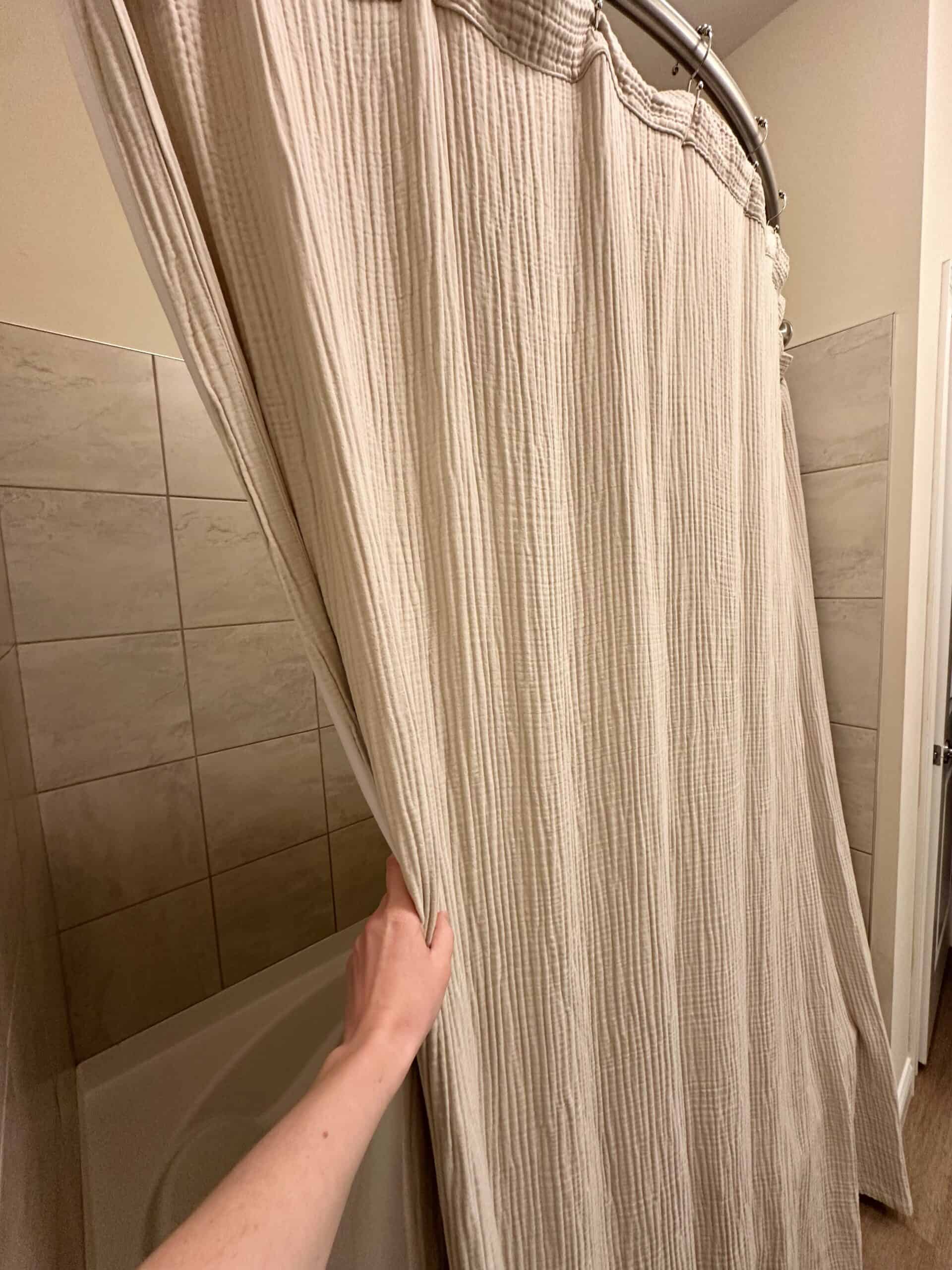 Hand pulling back a beige, textured cotton shower curtain in a tiled bathroom.