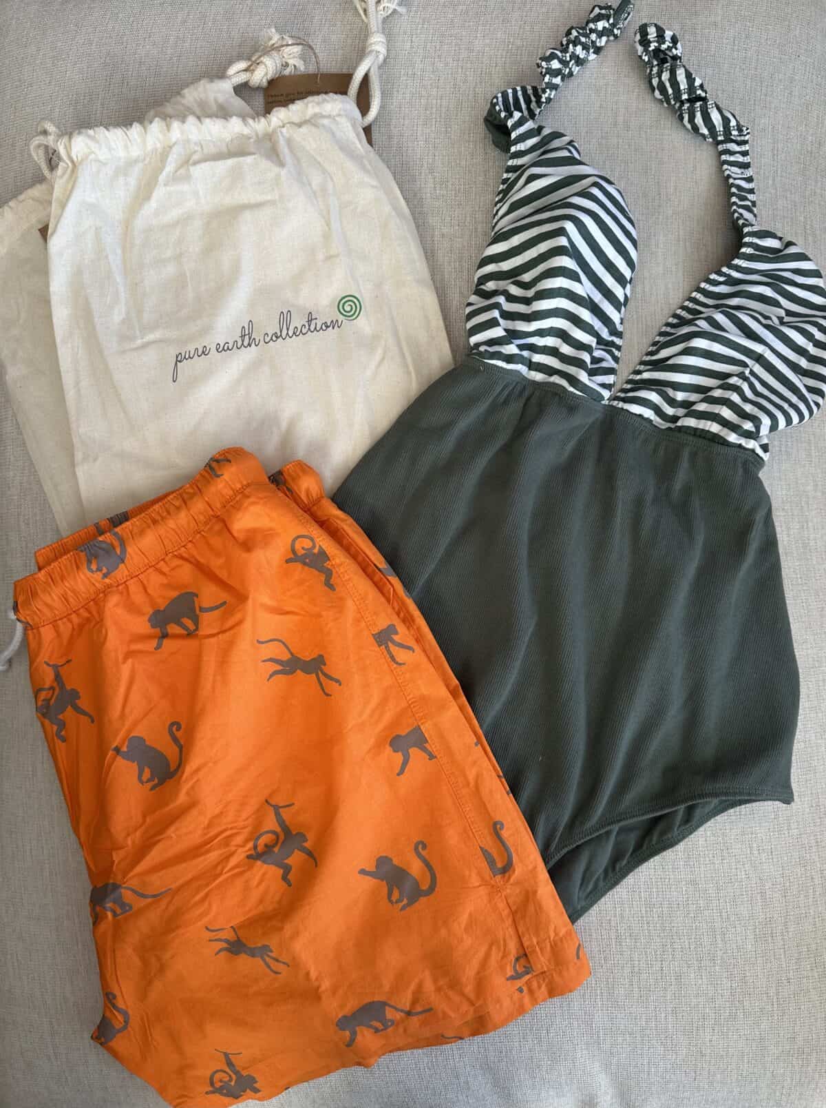 Two swimsuits—an orange pair of swim trunks with gray monkey prints and a green and white striped one-piece—laid out next to a drawstring bag labeled "pure earth collection."