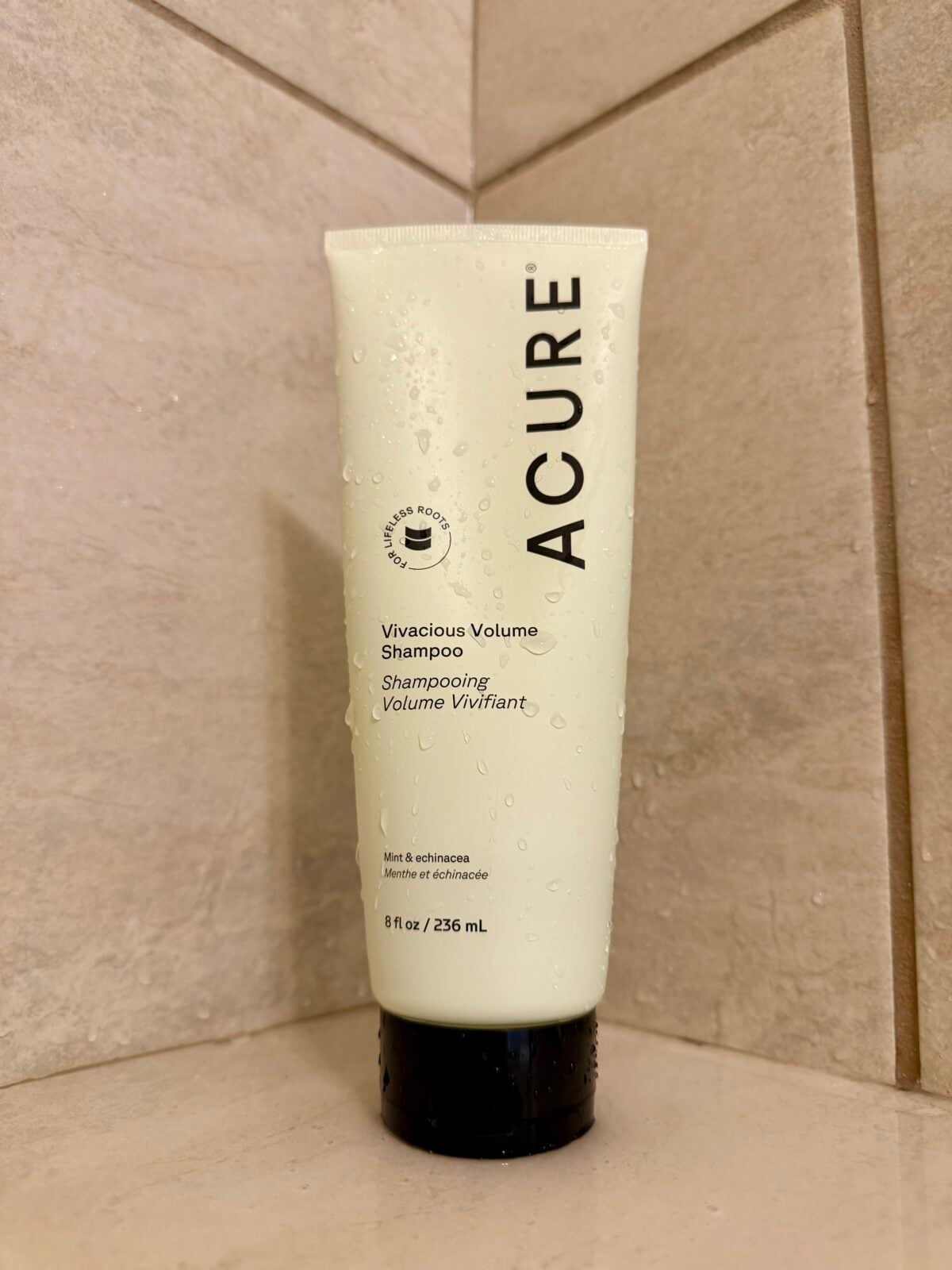 A beige tube of Acure Vivacious Volume Shampoo stands upright on a beige-tiled shower corner, with water droplets visible on the tube. The product label is in English and French. The tube contains 8 fl oz or 236 ml.