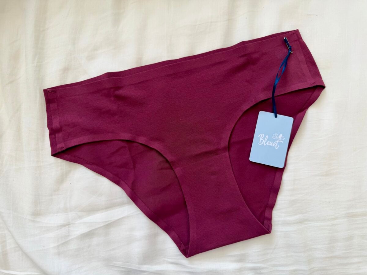 A pair of maroon-colored womens underwear with a blue tag labeled Bleuet attached, laid flat on a cream-colored fabric background.