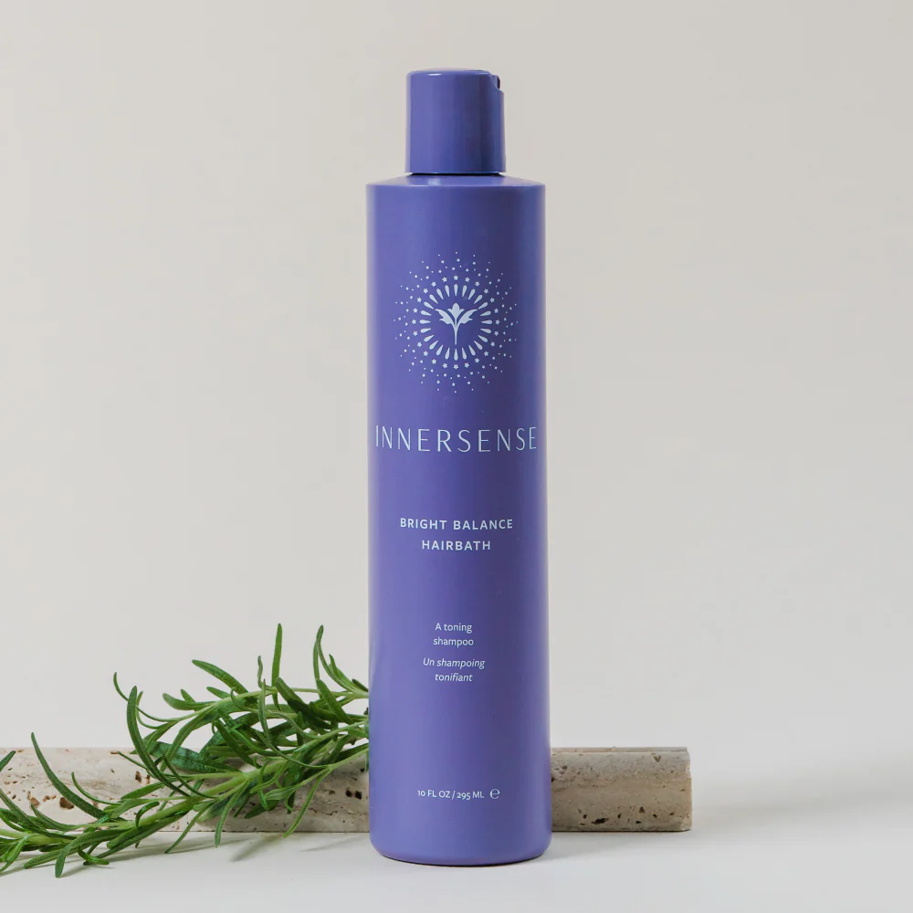 Purple bottle of Innersense Bright Balance Hairbath standing upright with a sprig of rosemary beside it on a light background.