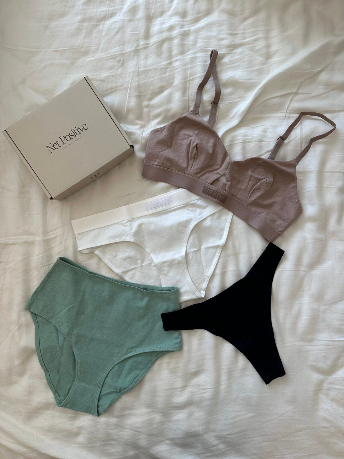 A box labeled Net Positive, a mauve bra, a green brief, a white brief, and a black thong are arranged on a white bedsheet.