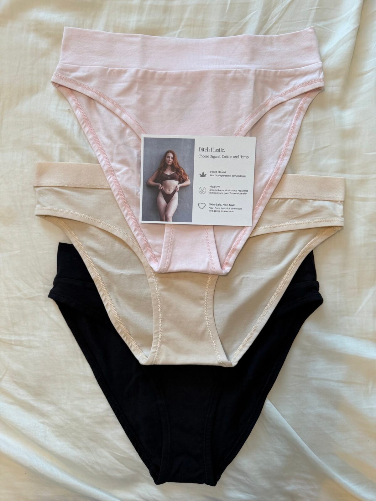 Three pairs of womens cotton underwear in pink, beige, and black are stacked on a white surface. A card with a photo and product information rests on the top, pink pair.