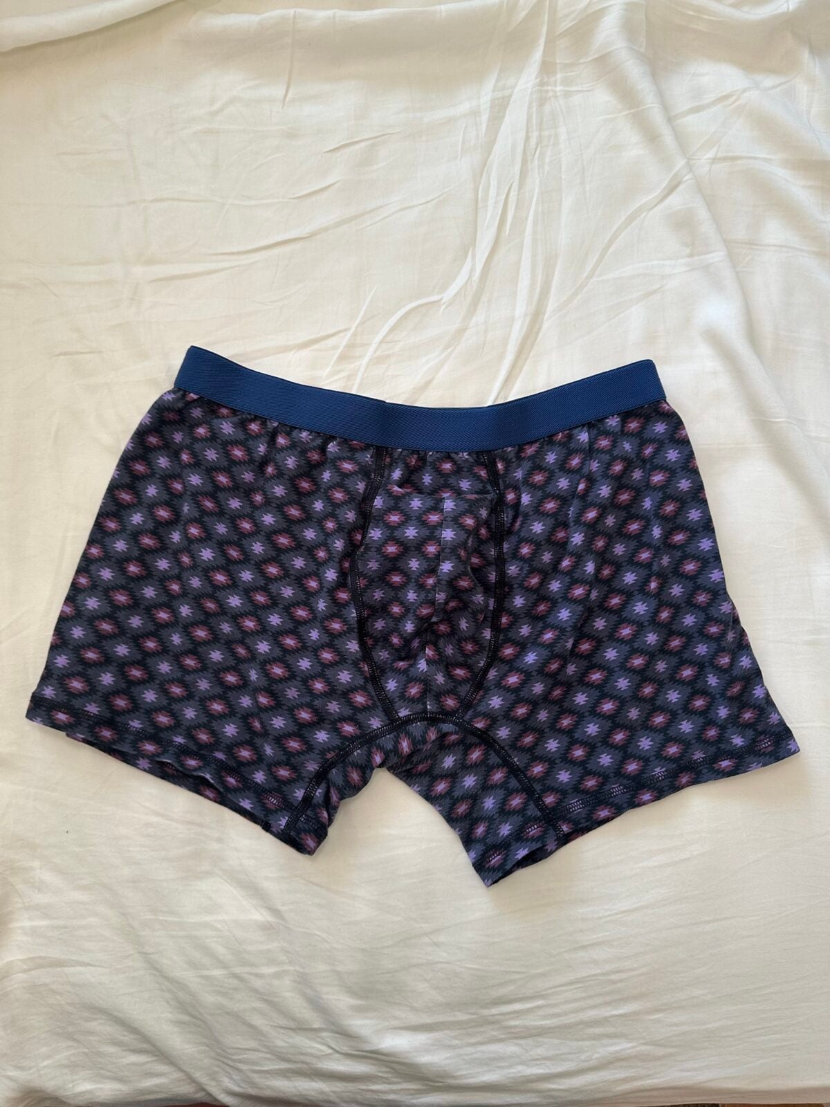 Men&rsquo;s patterned organic boxer briefs in dark blue and purple laid flat on a cream bed sheet.