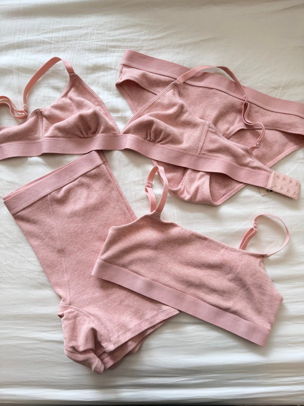 Four pieces of light pink underwear, including two bras, one pair of briefs, and one pair of high-waisted shorts, are neatly laid out on a white bedsheet.