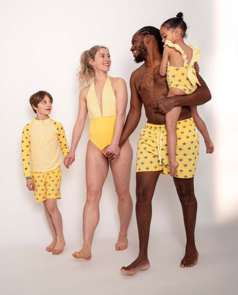 A family of four wearing matching yellow patterned swimwear poses against a white background, smiling and holding hands.