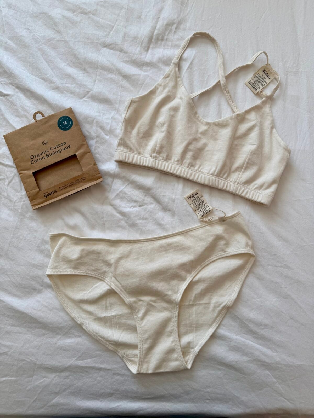 A matching set of off-white organic cotton underwear, consisting of a bralette and a pair of briefs, is laid out on a white surface. Tags are attached, and packaging is placed on the left.