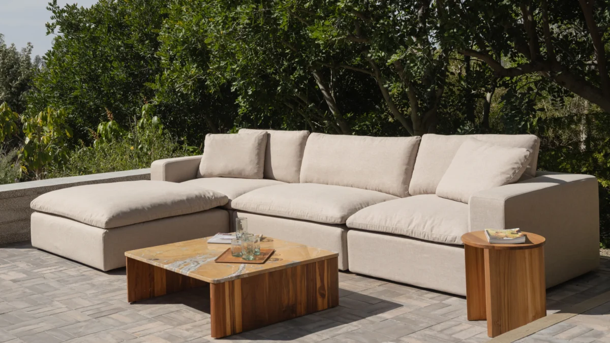 A beige sectional sofa with plush cushions is arranged outdoors on a stone patio, accompanied by a rectangular wooden coffee table and a small round wooden side table. Green trees and foliage are visible in the background.